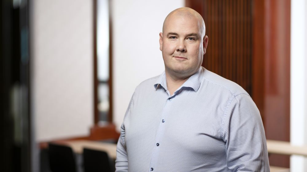 Kuvassa vienosti hymyilevä mies, jolla on sini-valkoinen pikkuruutuinen paitapusero.