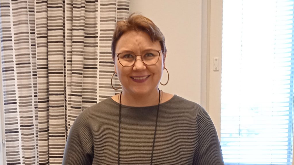 Nainen toimistotilassa. Hymyilee. Silmälasit, korvarenkaat ja khakin värinen paita.