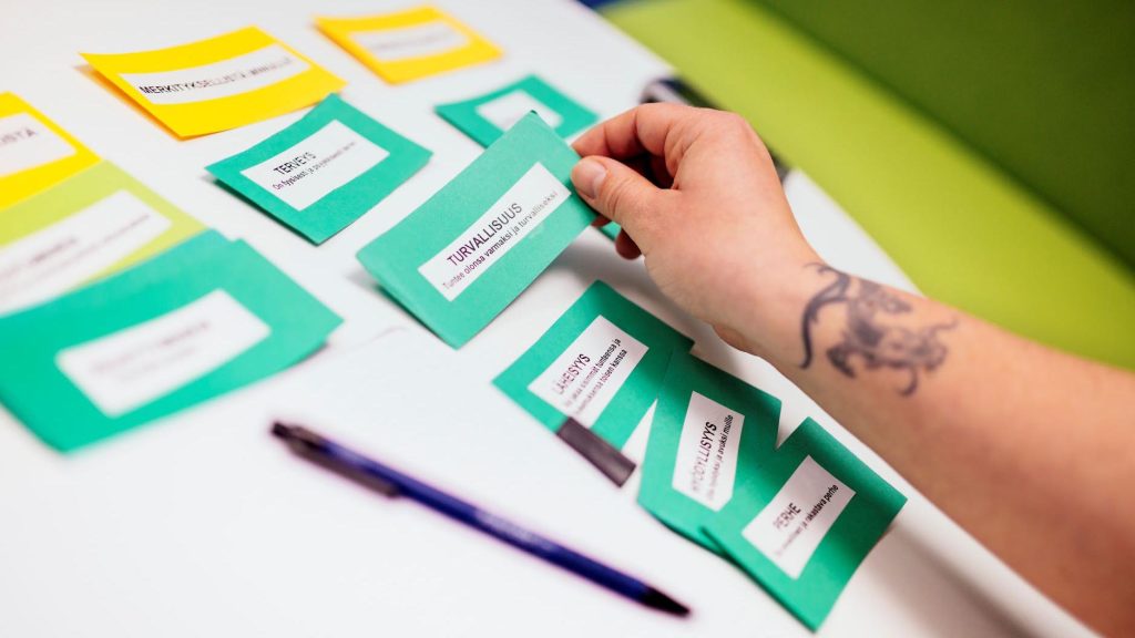 Pöy­däl­le on ase­tel­tu vä­rik­käi­tä lap­pu­ja. Ta­tuoi­tu käsi tart­tuu lap­puun, jossa lukee turvallisuus.