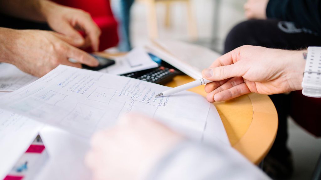 Työs­ken­te­lyä pöy­dän ää­rel­lä, jos­sa on kä­sin kir­joi­tet­tu­ja muis­tiin­pa­no­ja.