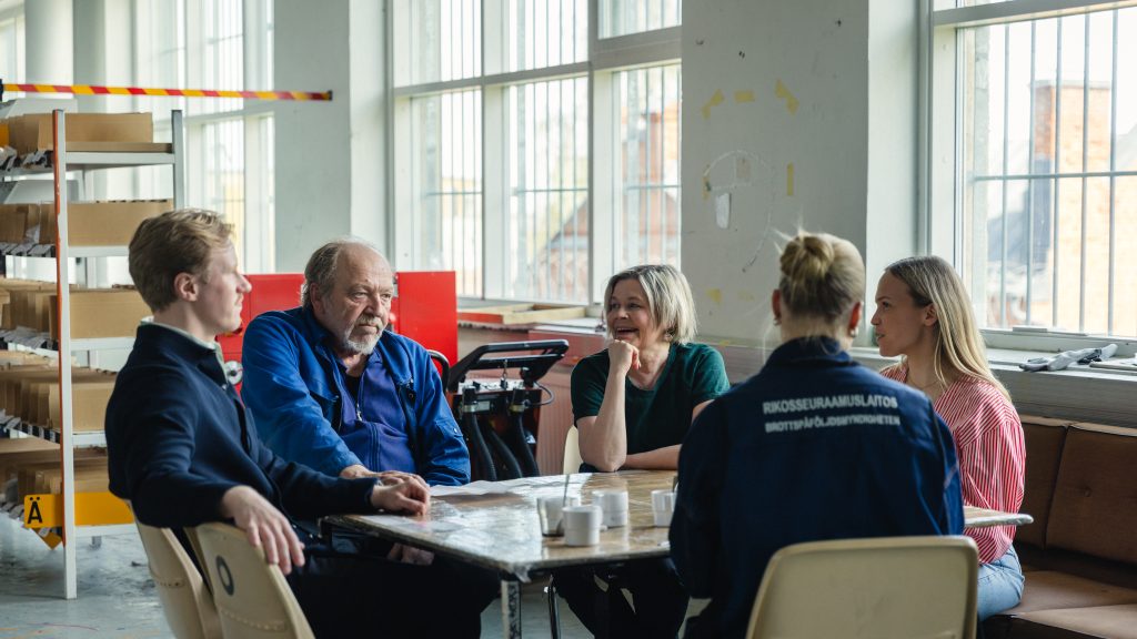 Teollisen näköisessä tilassa keskustelee viisi henkilöä. Kuvaan selin olevalla naisella on päällään työasu, jonka selkämyksessä lukee Rikosseuraamuslaitos.