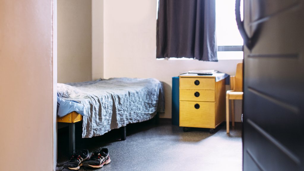 Vankilan selli. Näkyy sänky, lipasto ja pöytä sekä lenkkarit sängyn edessä.