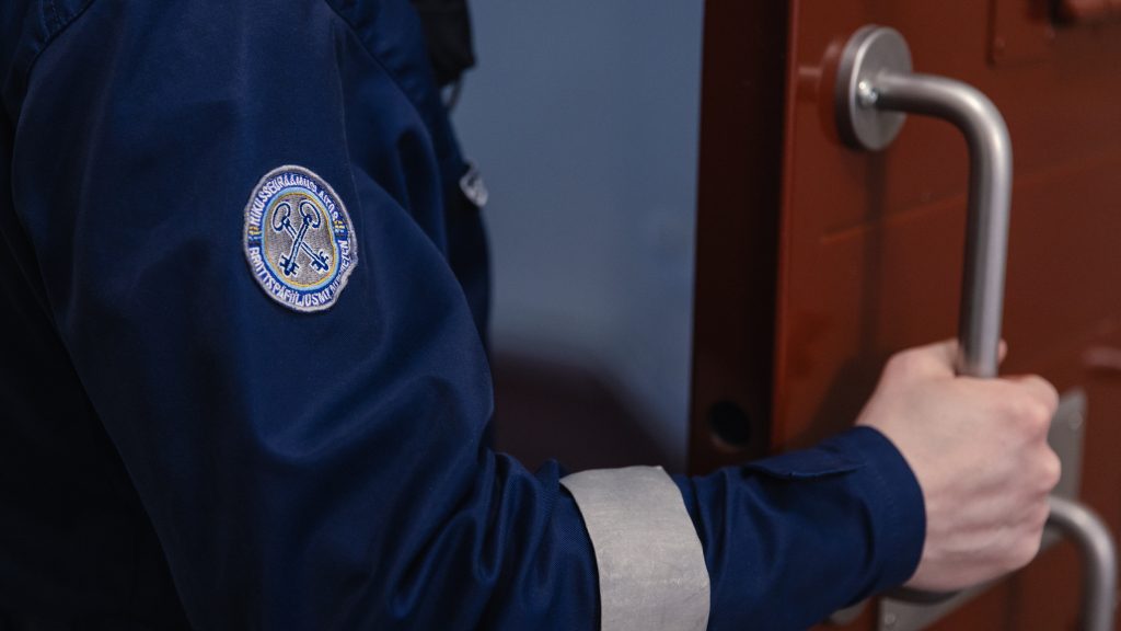 Rikosseuraamuslaitoksen työasuun pukeutunut henkilö pitää kädellään raskaan oven kahvasta.