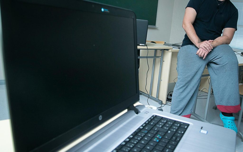 Luokkahuoneessa pulpetin päällä istuu henkilö collegehousuissa. Etualalla on kannettava tietokone.