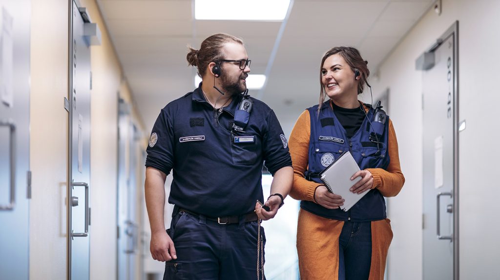 Kaksi Rikosseuraamuslaitoksen työasuun pukeutunutta henkilöä kulkee vankilan sellikäytävällä kohti kameraa ja keskustelee hymyillen.