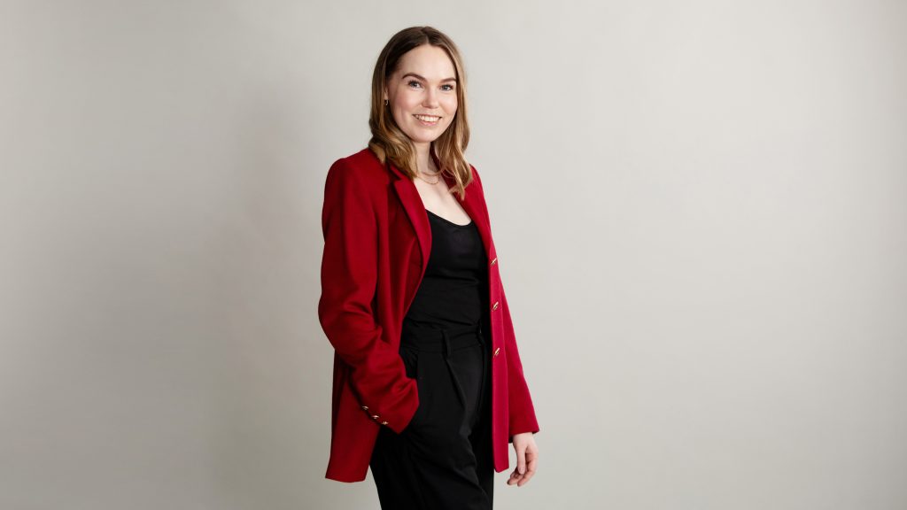 Kuvassa hymyilevä nainen, jolla on punainen jakku ja musta toppia ja mustat housut.