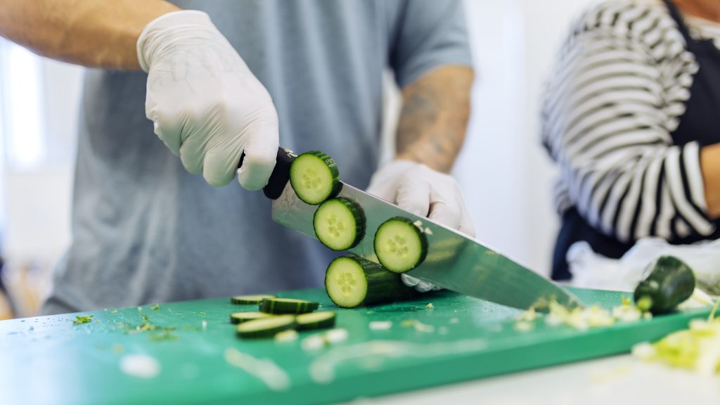 Henkilö leikkaa kurkkua. Vieressä näkyy toinen henkilö myös laittamassa ruokaa.