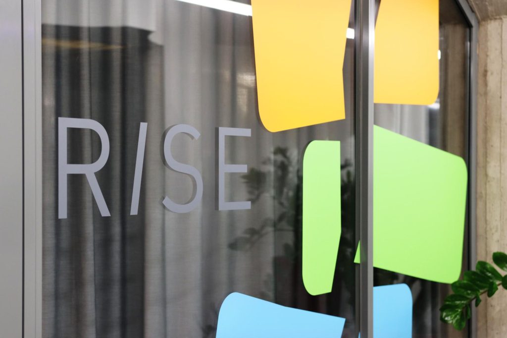 Logo ikkunalasissa. Logossa on teksti Rise ja erivärisiä graafisia kuvia.