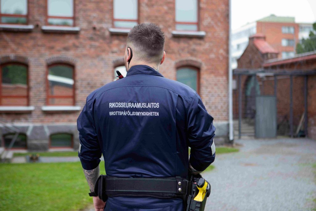 Vartija seisoo, takissa lukee Rikosseuraamuslaitos. Taustalla Vaasan vankila.