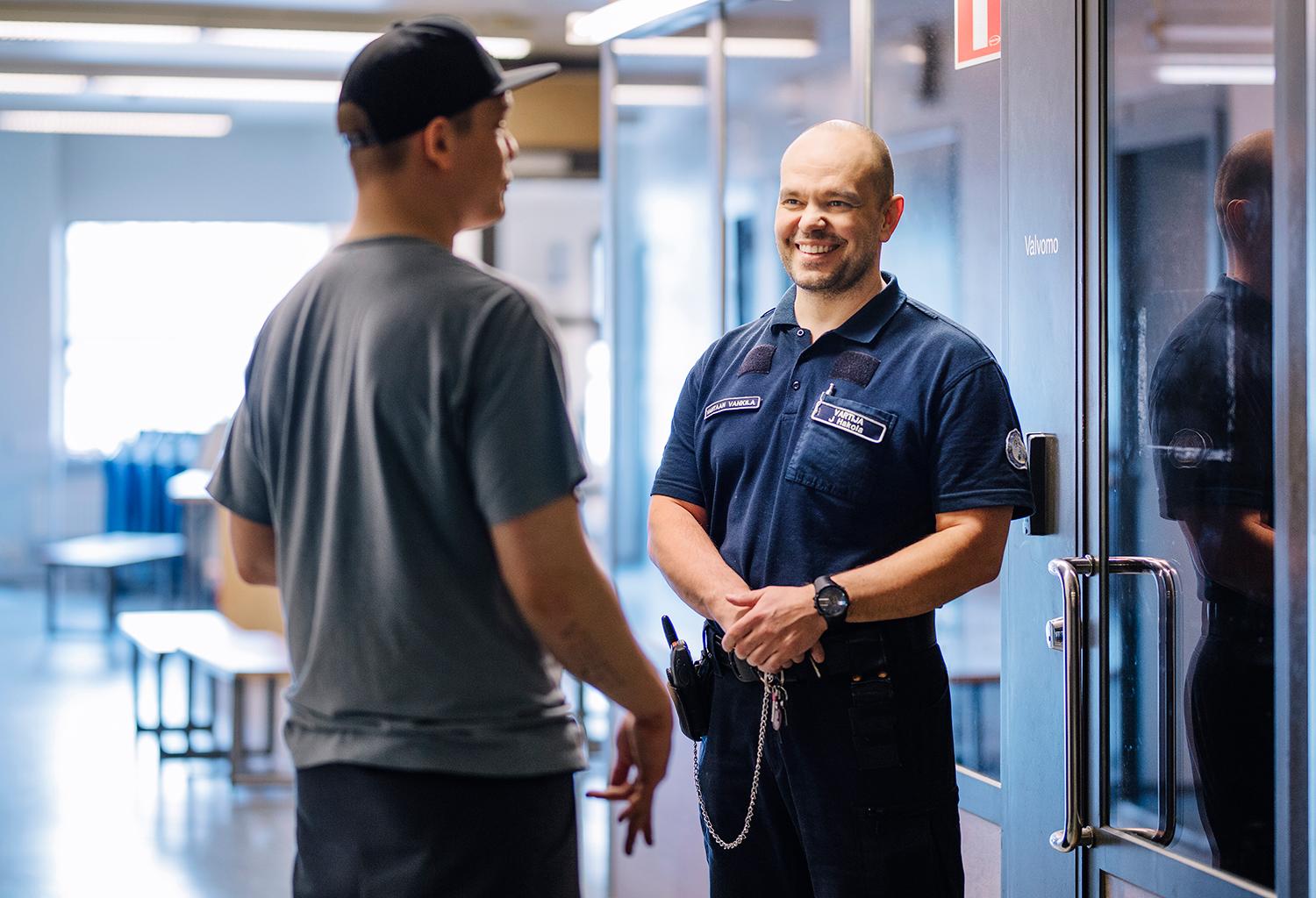 Two people are tal­king. One of them has his back to the ca­me­ra. The ot­her per­son facing the ca­me­ra is wea­ring a uni­form that says pri­son of­ficer on the front his shirt.