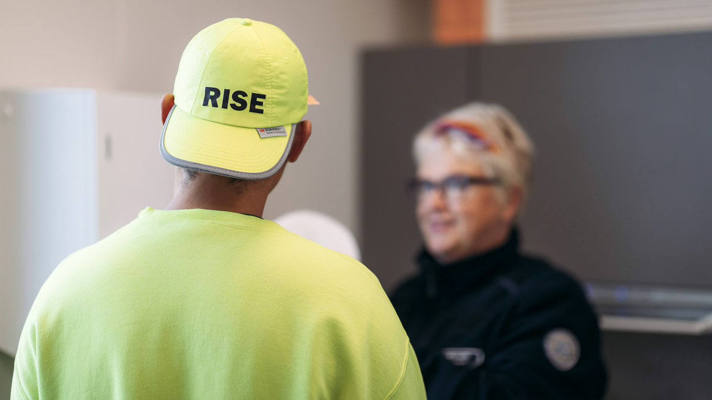 A man wearing a high-visibility shirt and a cap talks with an official of the Prison and Probation Service. The cap has the logo of the Prison and Probation Service on it.