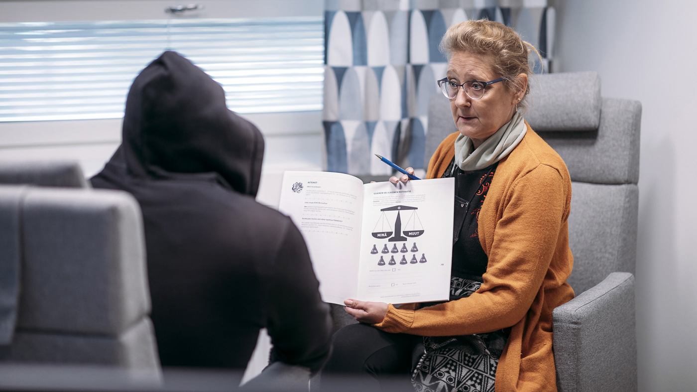 A woman shows a book to a person facing away from the camera.