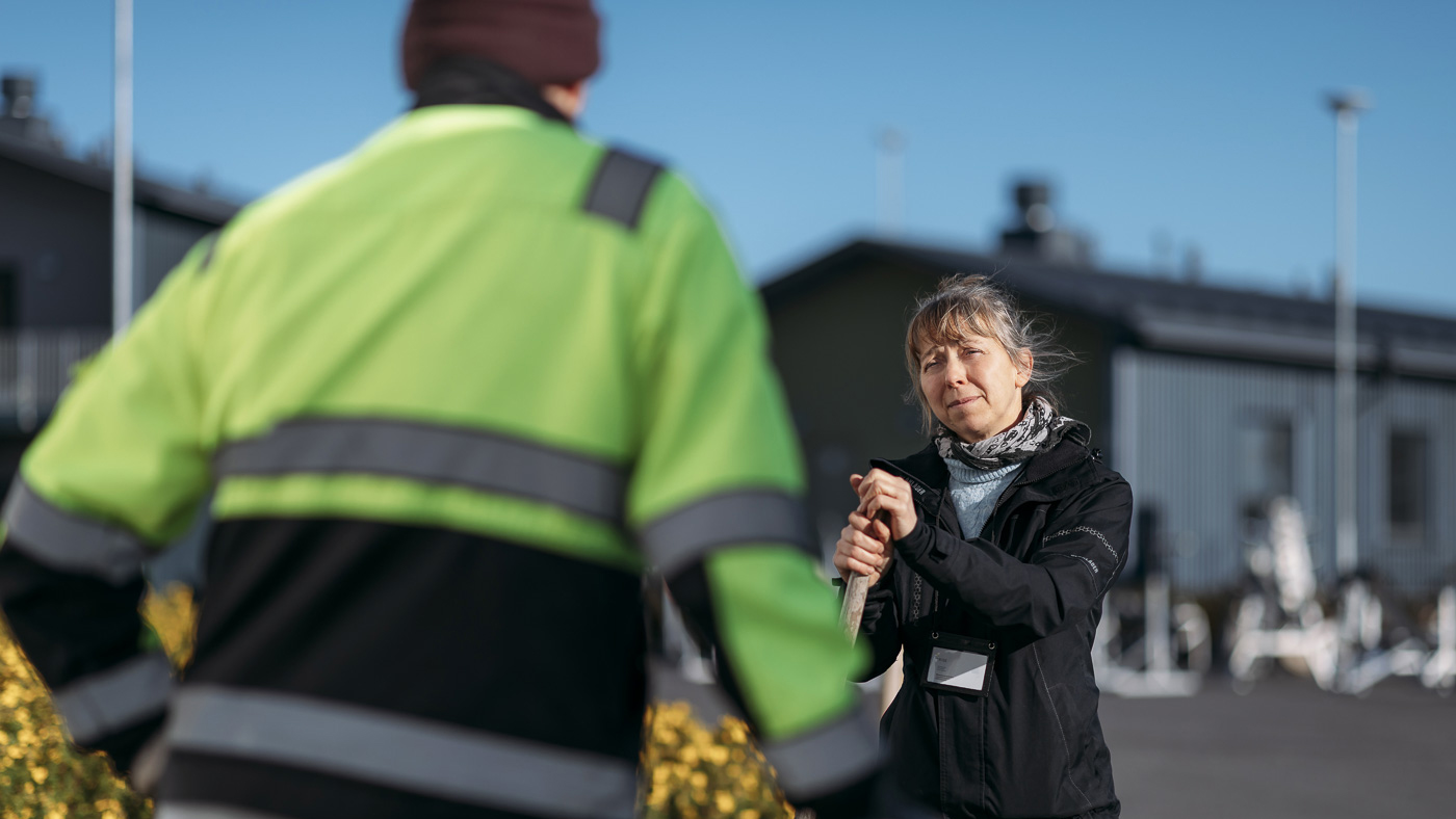 Huomioväriseen takkiin pukeutunut mies keskustelee naisen kanssa. Nainen nojaa harjanvarteen. Huomioväriseen takkiin pukeutunut mies keskustelee naisen kanssa. Nainen nojaa harjanvarteen.