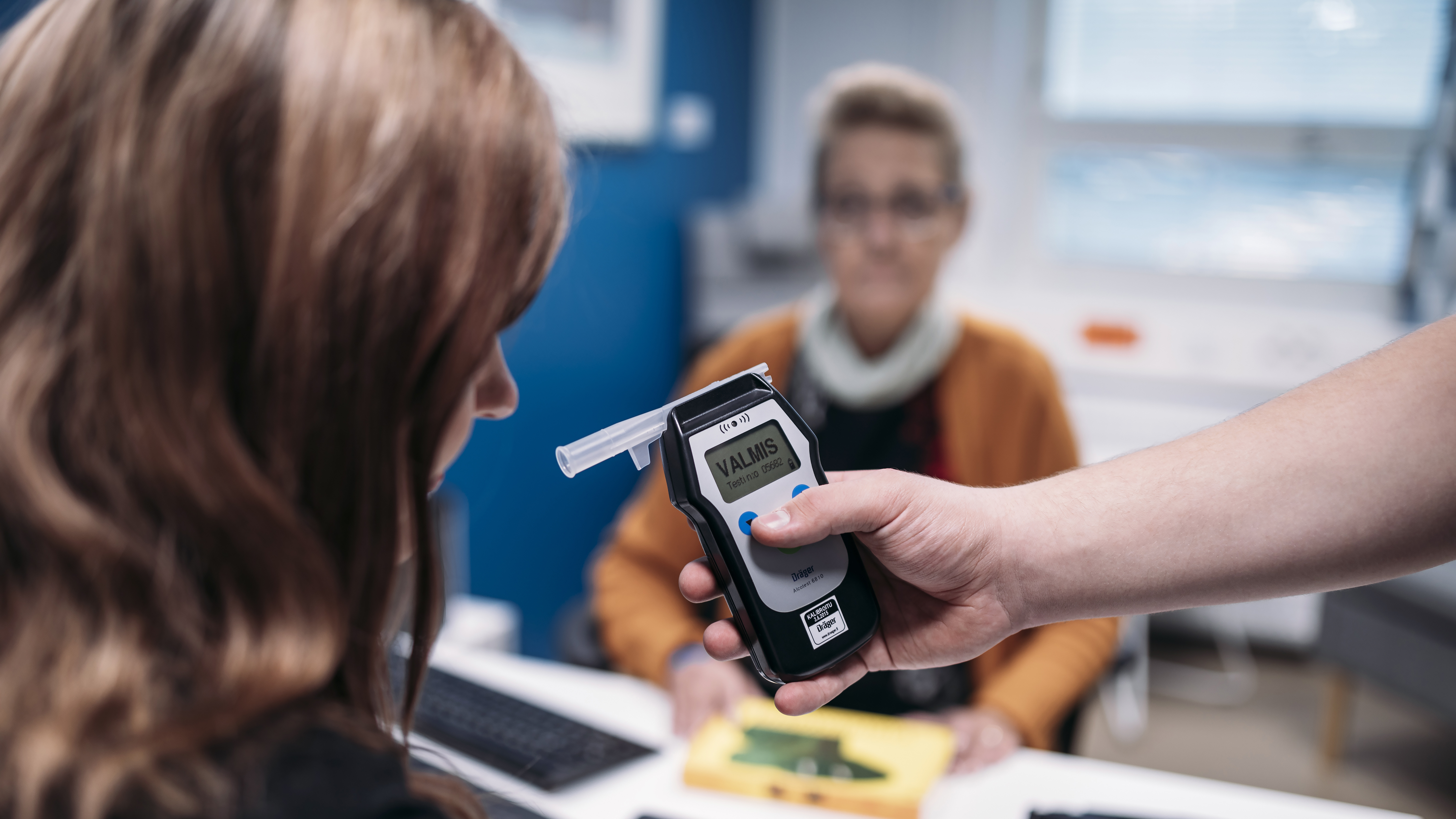 A person with long hair blows in a breathalyser as an official of the Prison and Probation Service watches in the background.
