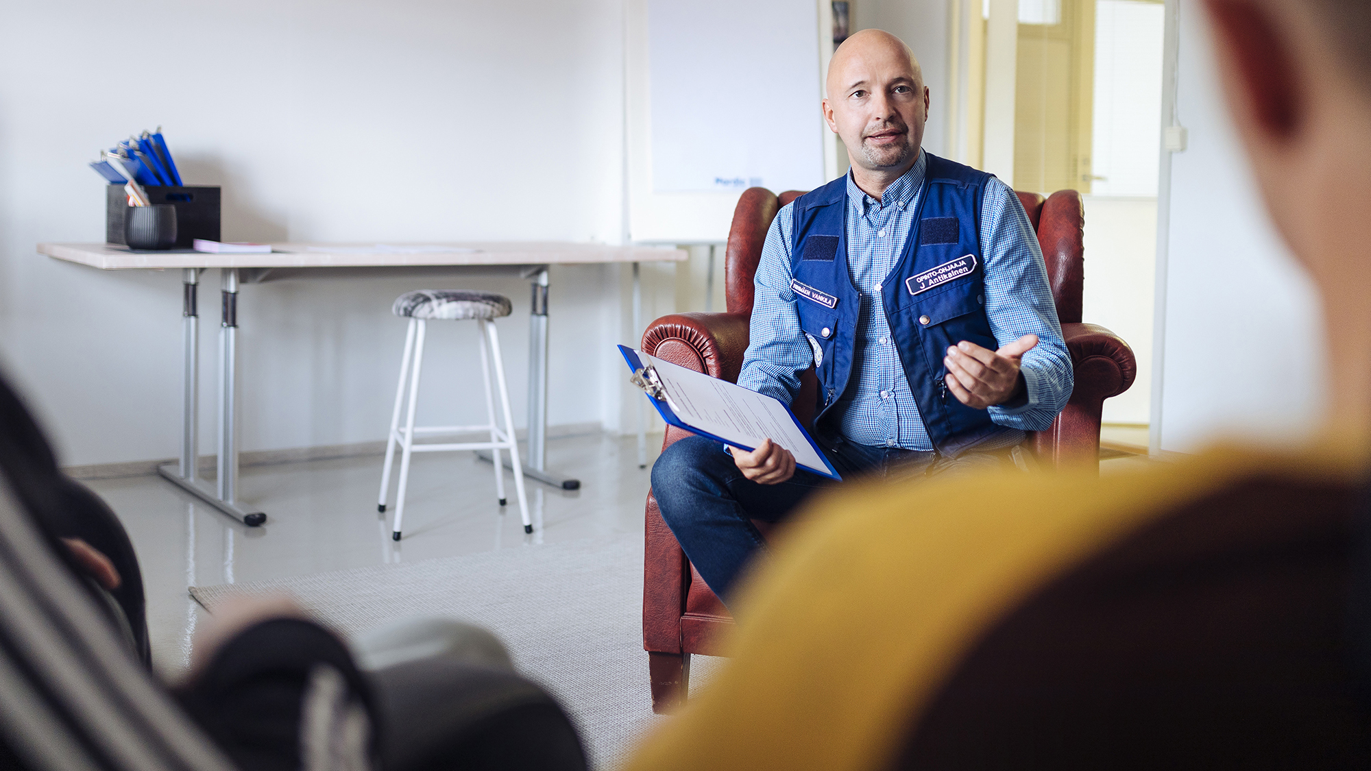 A man, who wears a multi-pocket vest with the Prison and Probation Service logo, sits in an armchair and talks with a people, who are facing away from the camera.