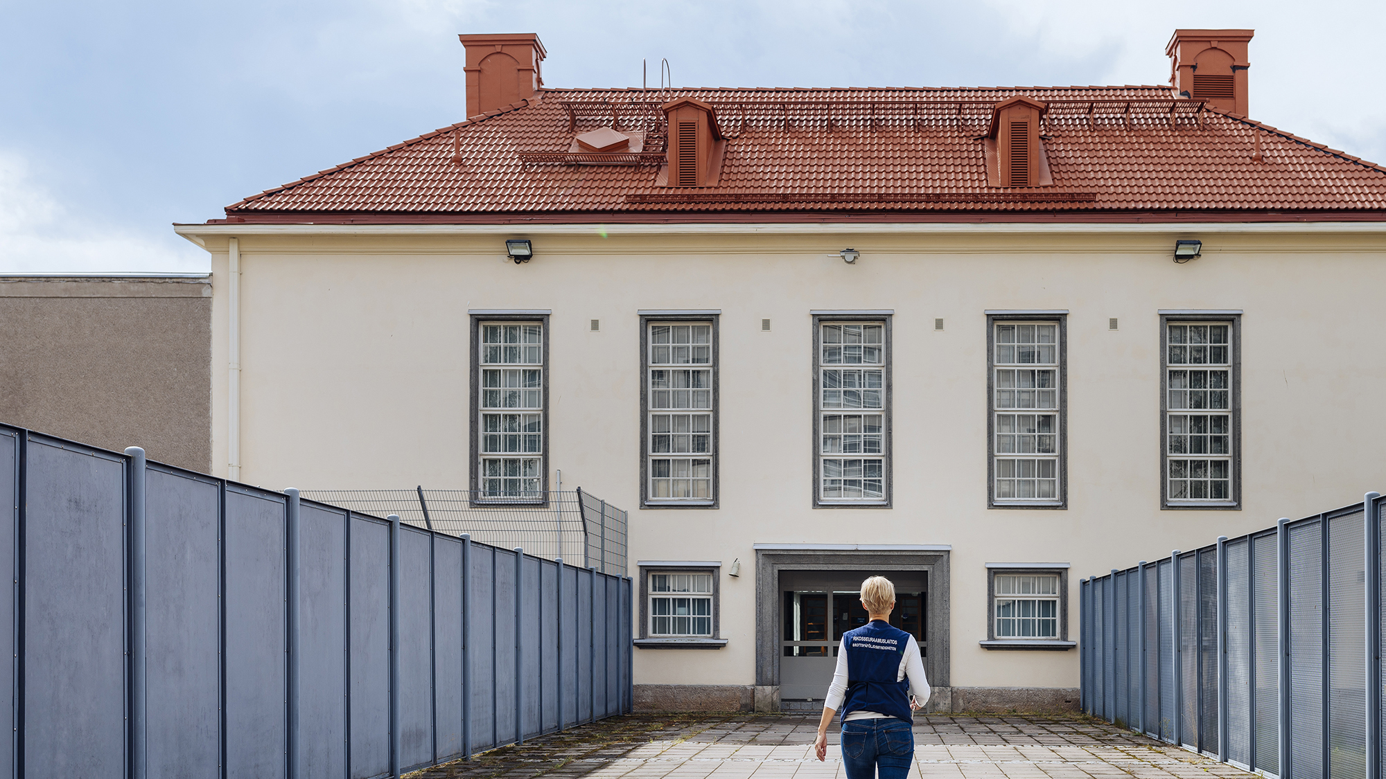 A person, who wears a multi-pocket vest with the Prison and Probation Service logo, walks towards a prison building.