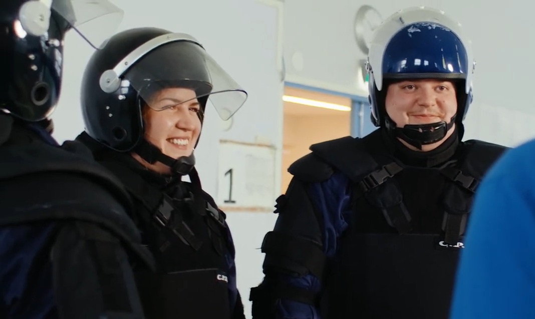 Två personer iklädda skottsäkra västar och skyddshjälmar samtalar med en tredje person som står med ryggen mot kameran. En av personerna som syns i foton har ett grin på ansiktet.